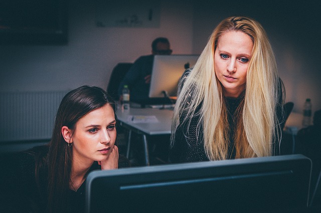 Two young, professional women working at a computer in an office room; one is sitting, the other is standing. 